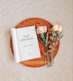 a book and some flowers on a wooden plate next to a white sheet that says love other words