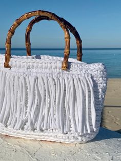 a white handbag sitting on top of a sandy beach next to the ocean with bamboo handles