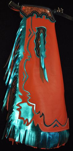 "Custom Chaps, handmade in the U.S.A.! These chaps are made with a quality, orange chap leather with metallic turquoise chap leather trim. They have 8 oz. tooling leather yokes, stained an antique Sheridan brown, with a black border and floral carved design on them. There is 10\" metallic turquoise and orange double fringe on the side and 4\" on the bottom. There are two chap snaps on each leg as well. These chaps are quality and flashy making them a nice choice for rodeo. Please message us for measurements when you order.  Thanks for looking! Note: We do custom work as well so If you need specific measurements or have something special in mind feel free to contact us. Our phone # is (406) 426-4300. We also have a variety of chinks, chaps and kid's chinks that are in stock now and ready fo Bull Riding Chaps, Custom Chaps, Western Chaps, Double Fringe, Riding Chaps, Bull Rider, Cowgirl Accessories, Bull Riders, Bull Riding