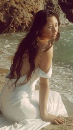 a woman sitting on top of a sandy beach next to the ocean with her hair blowing in the wind