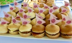 many small sandwiches and buns with pink flags on them are sitting on a white platter