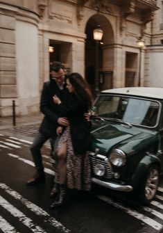 a man and woman standing next to a green car on the street in front of a building