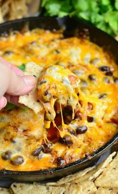 a hand holding a tortilla chip over a skillet filled with black beans and cheese