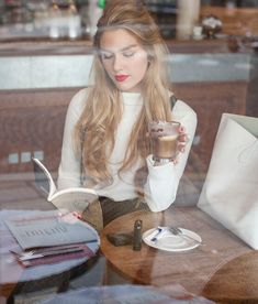 a woman sitting at a table reading a book and holding a drink in her hand