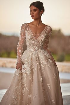 a woman wearing a wedding dress with long sleeves and flowers on the skirt is standing in front of an outdoor setting