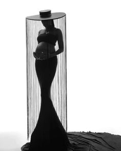 a pregnant woman standing in front of a tall glass vase with sheer curtains behind it