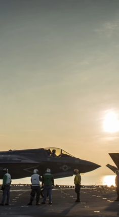 several people standing around a fighter jet on an aircraft carrier with the sun in the background