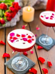 two glasses filled with drinks sitting on top of a wooden table next to red rose petals