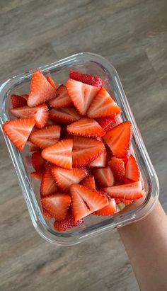 a person holding up a plastic container filled with sliced strawberries on top of a wooden table