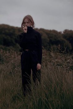 a woman standing in tall grass with her hand to her mouth and looking at the camera