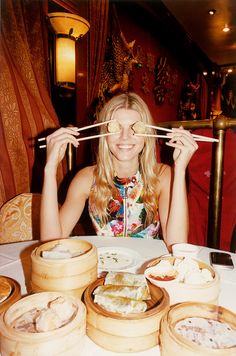 a woman holding chopsticks over her eyes while sitting at a table filled with food