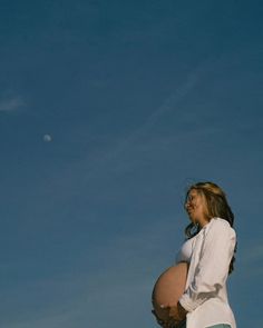 a pregnant woman standing under a blue sky with the moon visible in the back ground