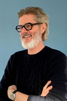 an older man with glasses and a beard smiles at the camera while standing in front of a blue background