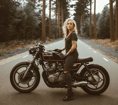 a woman is sitting on a motorcycle in the middle of the road with trees behind her