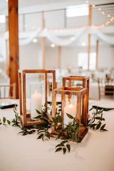 two lanterns with greenery and candles on top of a white table cloth at a wedding reception