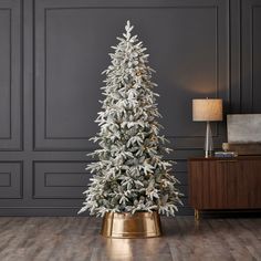 a white christmas tree in a room with wood flooring and grey walls, surrounded by a gold potted planter