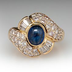 This elegant ring is centered with one (1), bezel set, oval cabochon cut natural sapphire. The sapphire is bordered with eight (8), channel set, tapered baguette cut diamonds and thirty-two (32), bead set, round brilliant cut diamonds. The ring measures 17.0mm at the top, rises 9.7mm above the finger, tapering to 3.8mm wide and 2.0mm thick at the base of the shank. This ring is currently a size 6. Formal Polished Finish Oval Cabochon Sapphire Ring, Classic Cabochon Sapphire Ring For Formal Occasions, Classic Formal Cabochon Sapphire Ring, Formal Oval Cabochon Sapphire Ring, Classic Formal Sapphire Cabochon Ring, Formal Sapphire Ring With Bezel Setting And Oval Cabochon, Elegant Cabochon Sapphire Ring, Formal Fine Jewelry Sapphire Ring Oval Cabochon, Elegant Domed Sapphire Ring In Yellow Gold