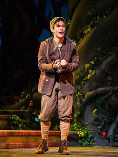 a man standing on top of a stage wearing brown pants and a hat with his hands in his pockets