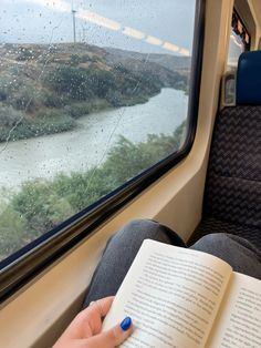 a person sitting on a train reading a book and looking out the window at a river