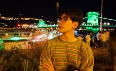 a young man standing in front of a city at night with lights on the bridge behind him