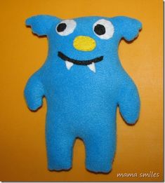 a blue stuffed animal with big eyes and a smile on it's face, standing against a yellow wall