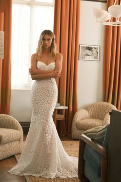 a woman in a white wedding dress standing in front of a window with her arms crossed