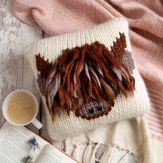 a cup of coffee and an open book on top of a table next to a knitted pillow