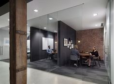 two people are sitting at a table in an office with glass walls and brick wall