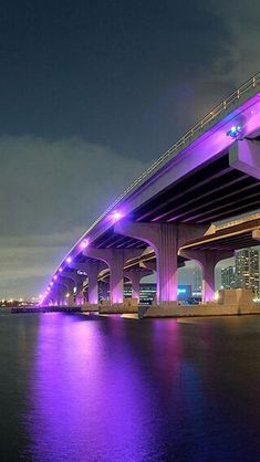 the bridge is lit up at night with purple lights on it's sides and reflecting in the water