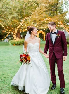 a bride and groom hold hands as they walk through the grass with lights in the background