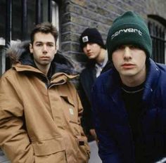 three young men standing next to each other in front of a brick building, one wearing a green beanie