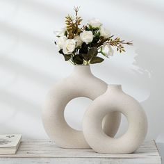 two white vases with flowers in them sitting on a table next to a book