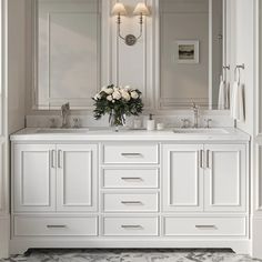 a white bathroom with two sinks, mirrors and flowers on the counter top in front of it