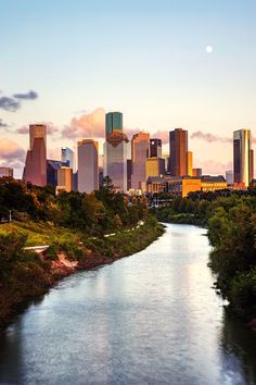 a river running through a lush green forest next to a tall cityscape at sunset