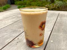 a glass filled with liquid sitting on top of a wooden table next to grass and bushes