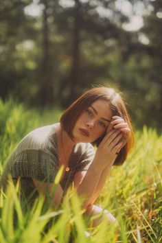 a woman is sitting in the grass with her hands on her head and looking at the camera