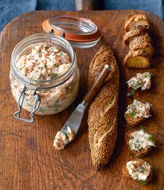 a wooden cutting board topped with bread next to a jar of dip and other food