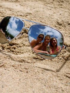 two people taking a selfie in the reflection of their sunglasses on the sand at the beach