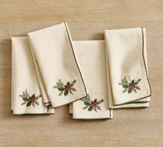 four christmas holly embroidered napkins on top of a wooden table with red berries and green leaves