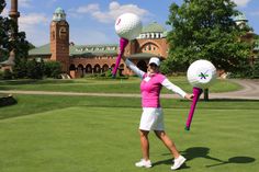 a woman in pink shirt and white skirt holding golf balls on green field next to large building