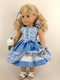 a doll with blonde hair wearing a blue dress and white shoes, holding a flower in her hand