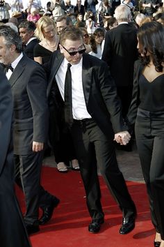 two people in suits and sunglasses walking on a red carpet with other people behind them