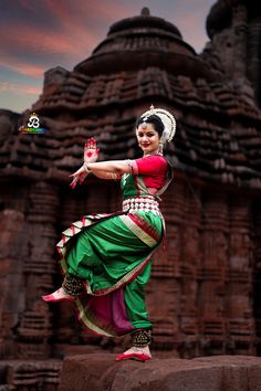 a woman in a green and red dress is dancing on a stone platform with an ornate structure behind her