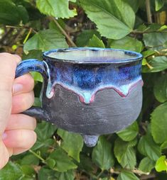 a hand holding a blue and black coffee mug in front of some green leaves on a sunny day