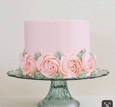 a cake with pink frosting and roses on it sitting on a glass stand against a white wall