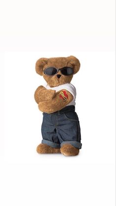 a brown teddy bear wearing sunglasses and denim shorts, standing with his arms folded in the air