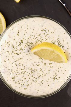 a glass bowl filled with white sauce next to lemons and herbs on a black surface