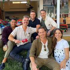 a group of men and women sitting on the grass in front of a house smiling at the camera