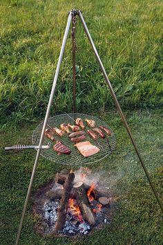 a grill with meat on it sitting in the grass next to a campfire and some tongs