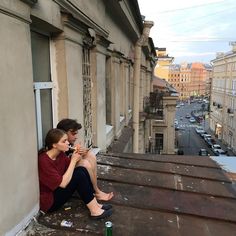 two people sitting on the roof of an old building looking at their cell phones and drinking beer